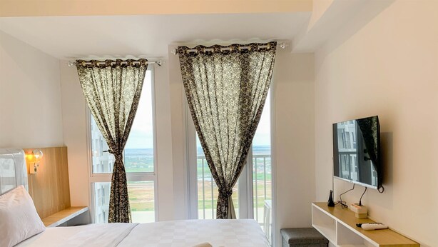A hotel room with a large bed, two windows with patterned curtains, a TV on the wall, and a small wooden shelf with items. - Restful And Cozy Studio Tokyo Riverside Pik 2 Apartment (Teluknaga)