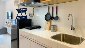 A modern kitchen with white tiles, a stainless steel sink, wooden shelves, and various kitchen utensils. - Restful And Cozy Studio Tokyo Riverside Pik 2 Apartment (Teluknaga)