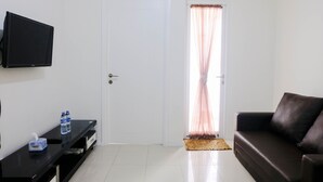 A living room with a black sofa, a flat-screen TV mounted on the wall, a small table with a tissue box and water bottles, and a door with sheer curtains. - Good Deal 1Br Apartment At Parahyangan Residence (Bandung)