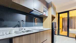 Una cocina moderna con un lavamanos, grifo de acero inoxidable y armarios de madera. Se observa un refrigerador a la derecha y un acceso a una sala de estar.