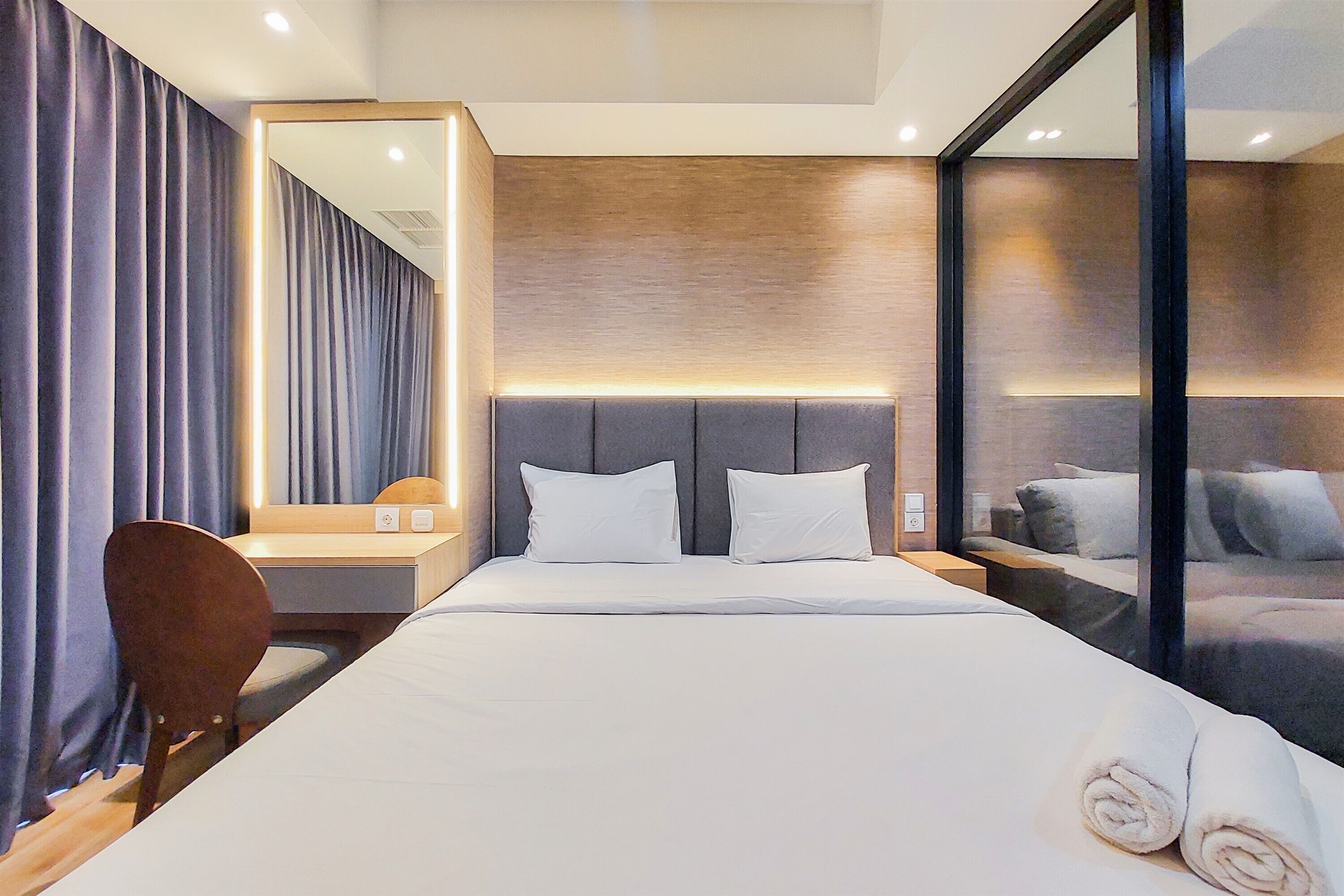 A modern hotel room with a large bed, a desk, a chair, a mirror, and a view of the city through a glass wall.