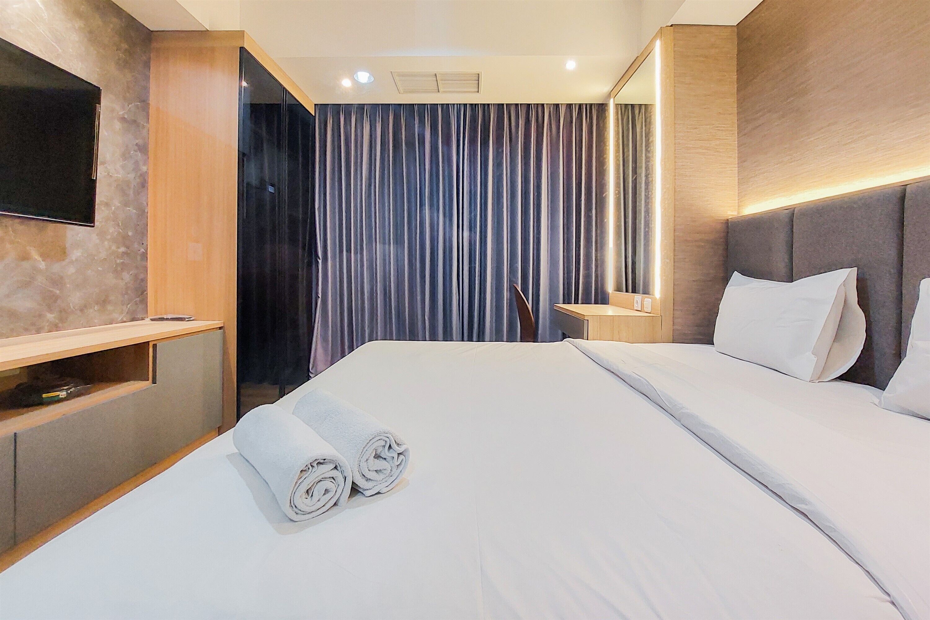 A hotel room with a neatly made bed, a flat-screen TV mounted on the wall, a wooden nightstand, and a large window with curtains.