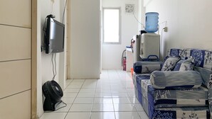 A room with a TV mounted on the wall, a water dispenser, a sofa with patterned cushions, and a tiled floor. - Cozy 2Br At Delta Cakung Apartment (Jakarta)