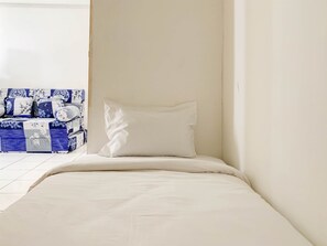 Una cama pequeña y bien hecha con un almohadón blanco y un edredón blanco en una habitación con paredes blancas y suelo de baldosas.