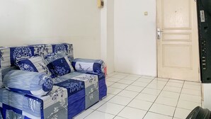 A room with a sofa covered in blue and white floral upholstery, a closed door, and a tiled floor. - Cozy 2Br At Delta Cakung Apartment (Jakarta)