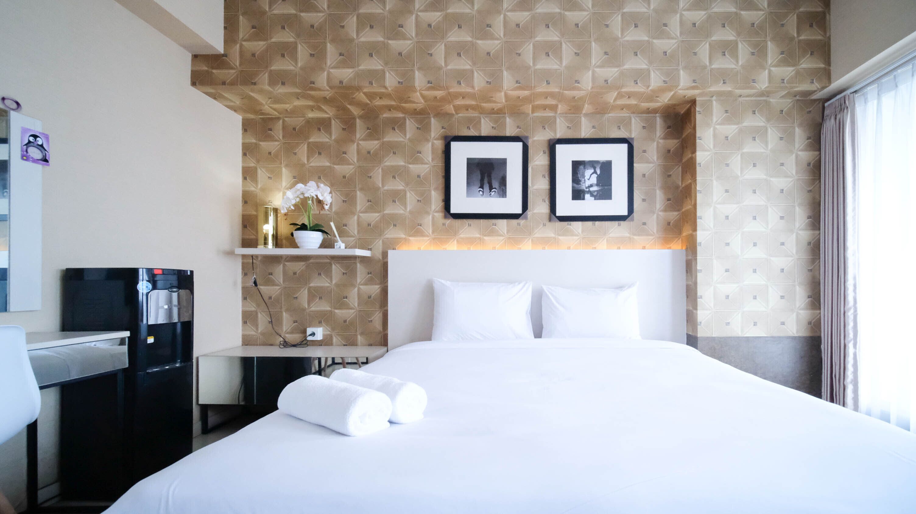 A hotel room with a large bed, a desk with a coffee machine, a small shelf with a plant, and two framed pictures on the wall.