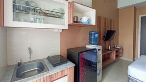 A compact kitchen with a built-in dishwasher, a water dispenser, and a flat-screen TV on a wooden shelf. - Classic Studio At Student Castle Yogyakarta Apartment (Depok)