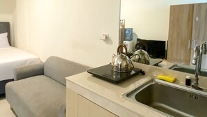 A compact hotel room with a bed, a grey sofa, a kitchenette with a kettle, a water dispenser, and a sink.