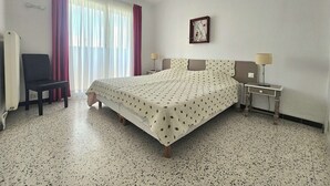 Ein Hotelzimmer mit Bett, Sessel, Nachttisch, Lampe, Bilderrahmen und einem Fenster mit Vorhängen.