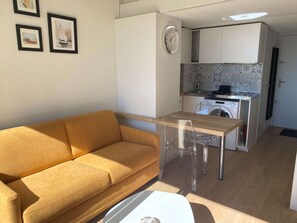 Un espace de vie compact comprenant un canapé, une table à manger avec des chaises, un coin cuisine avec un lave-linge et une horloge au mur.