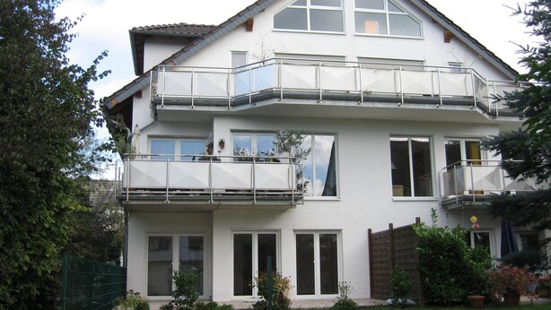 Un immeuble résidentiel de deux étages, doté d’un balcon à l’étage supérieur, d’un petit jardin à l’avant et d’une clôture en bois qui entoure la propriété.