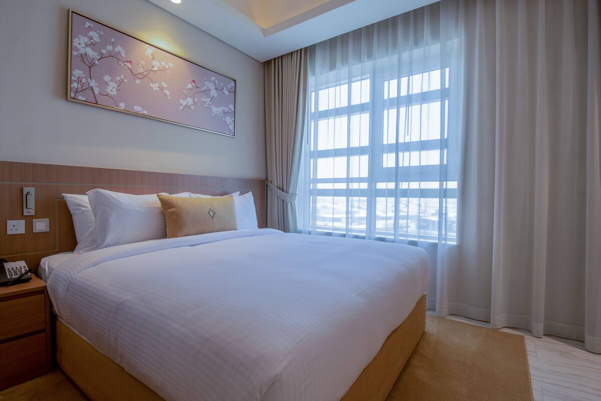 Ein Hotelzimmer mit einem großen Bett, einem Nachttischtelefon, einem an der Wand befestigten Lichtschalter und einem Fenster mit durchsichtigen Vorhängen.