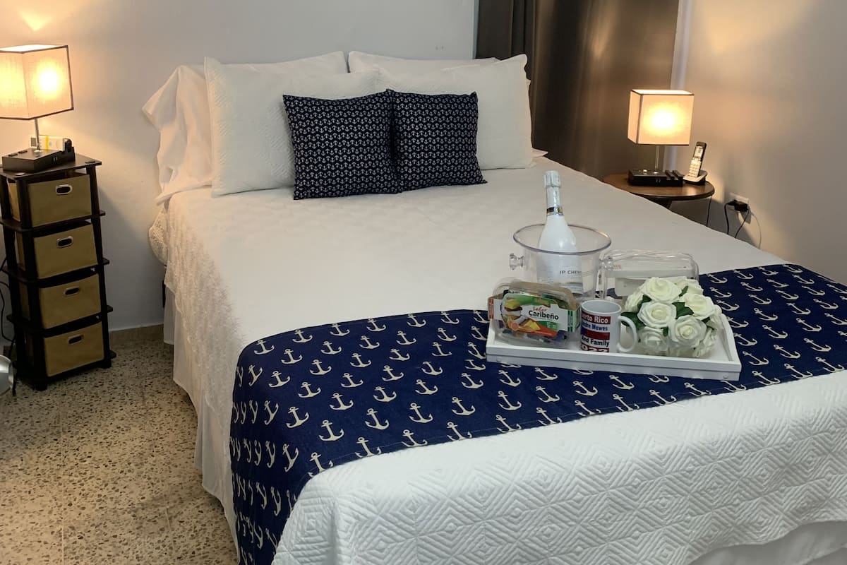 Habitación de hotel ordenada con una cama, dos mesitas de noche con lámparas, una bandeja con artículos de desayuno y un equipo de aire acondicionado en la pared.