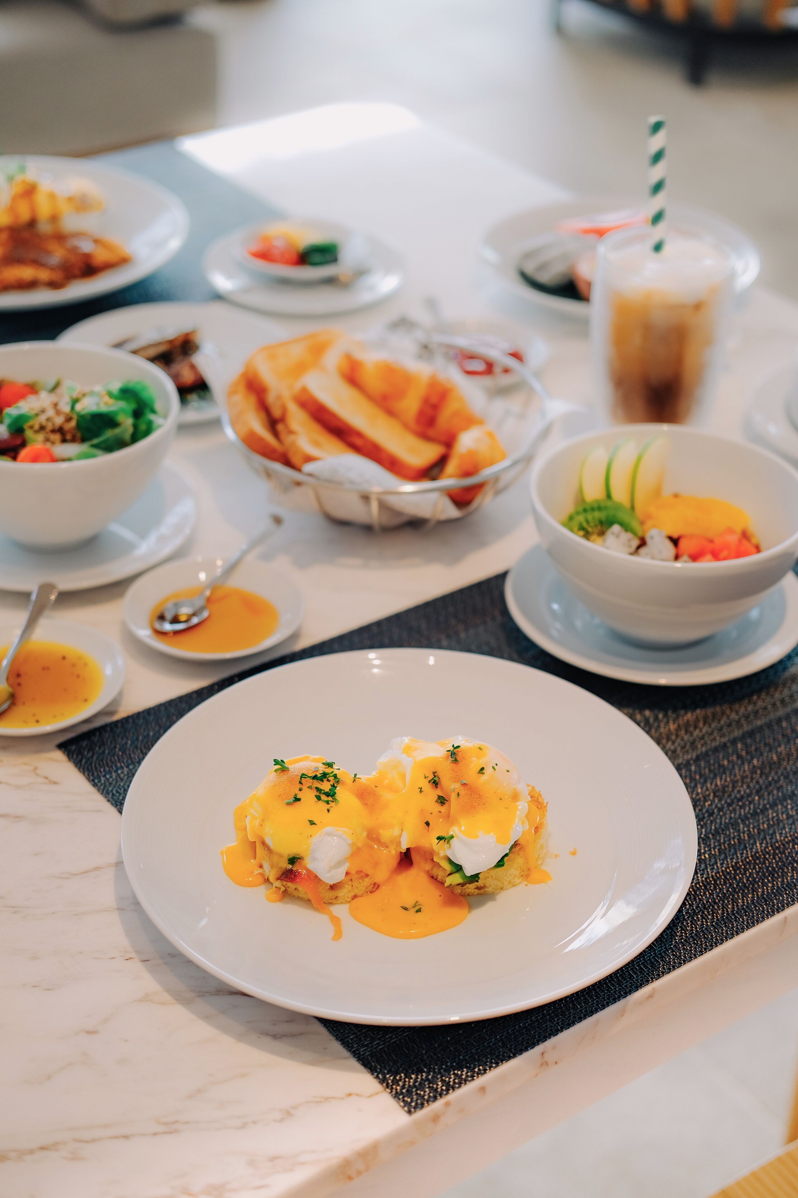 Una mesa de comedor con varios platos, incluyendo un plato de huevos Benedictinos, un tazón de ensalada y un vaso de café helado.