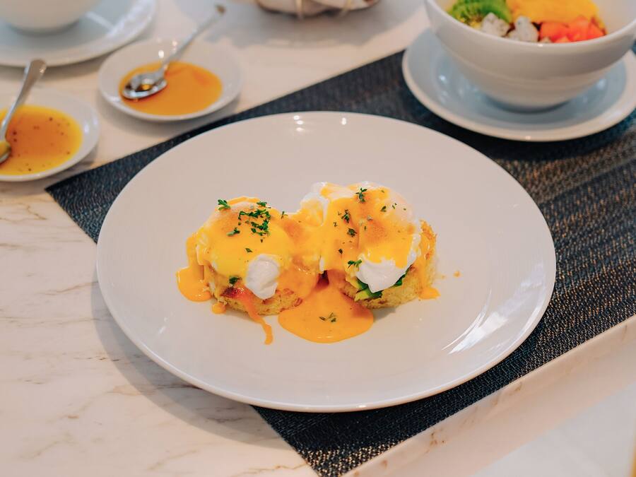 Una mesa de comedor con varios platos, incluyendo un plato de huevos Benedictinos, un tazón de ensalada y un vaso de café helado.
