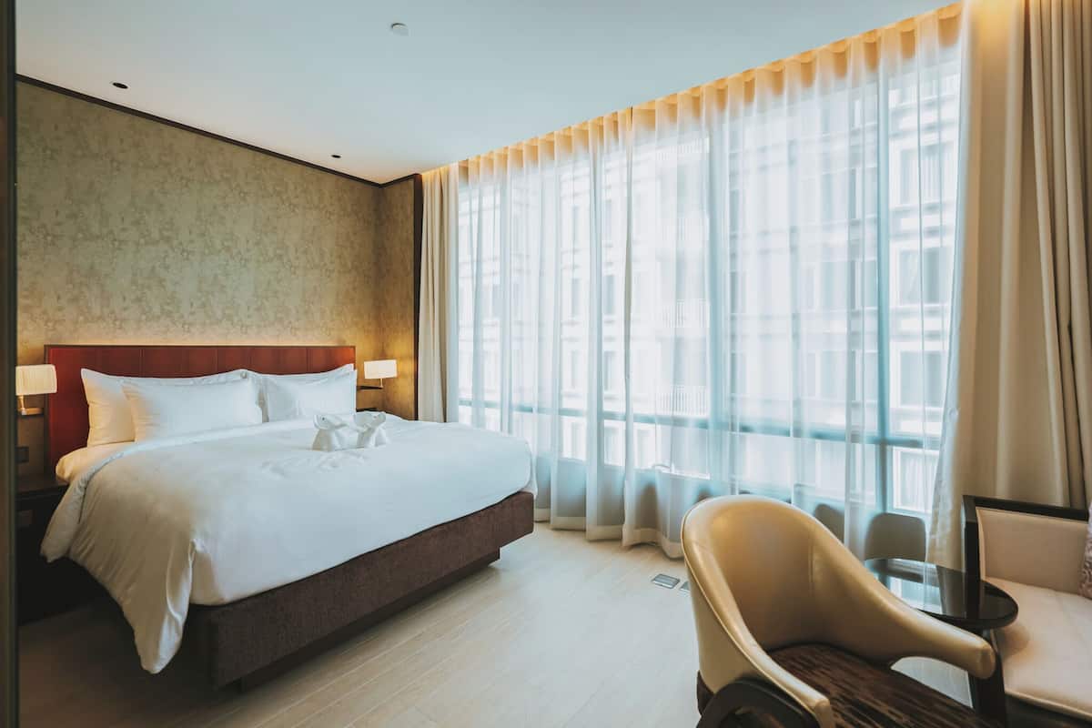 A hotel room with a large bed, a chair, and a view of the city through sheer curtains.