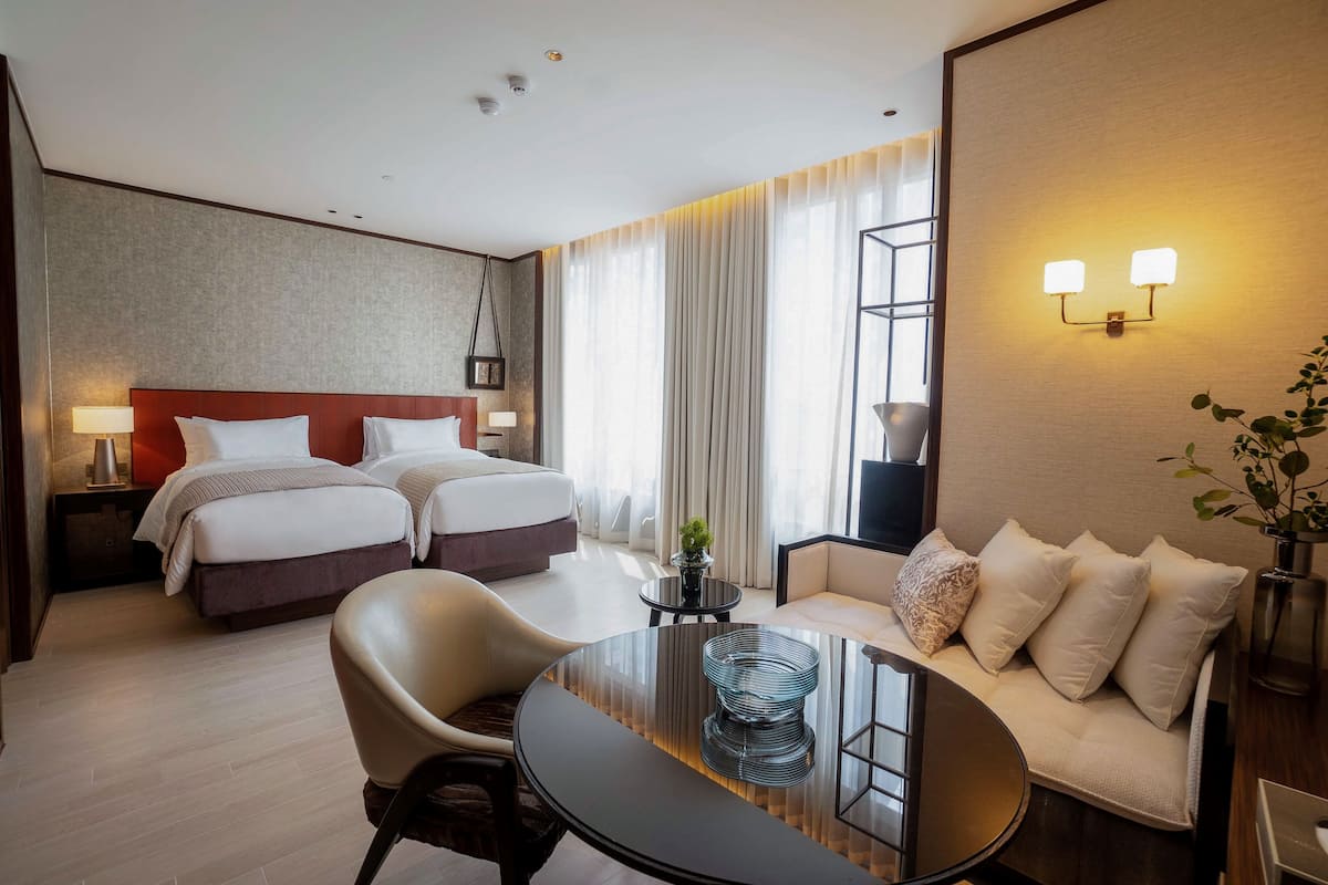 A hotel room with two beds, a sofa, a dining table, and a small side table with a lamp.