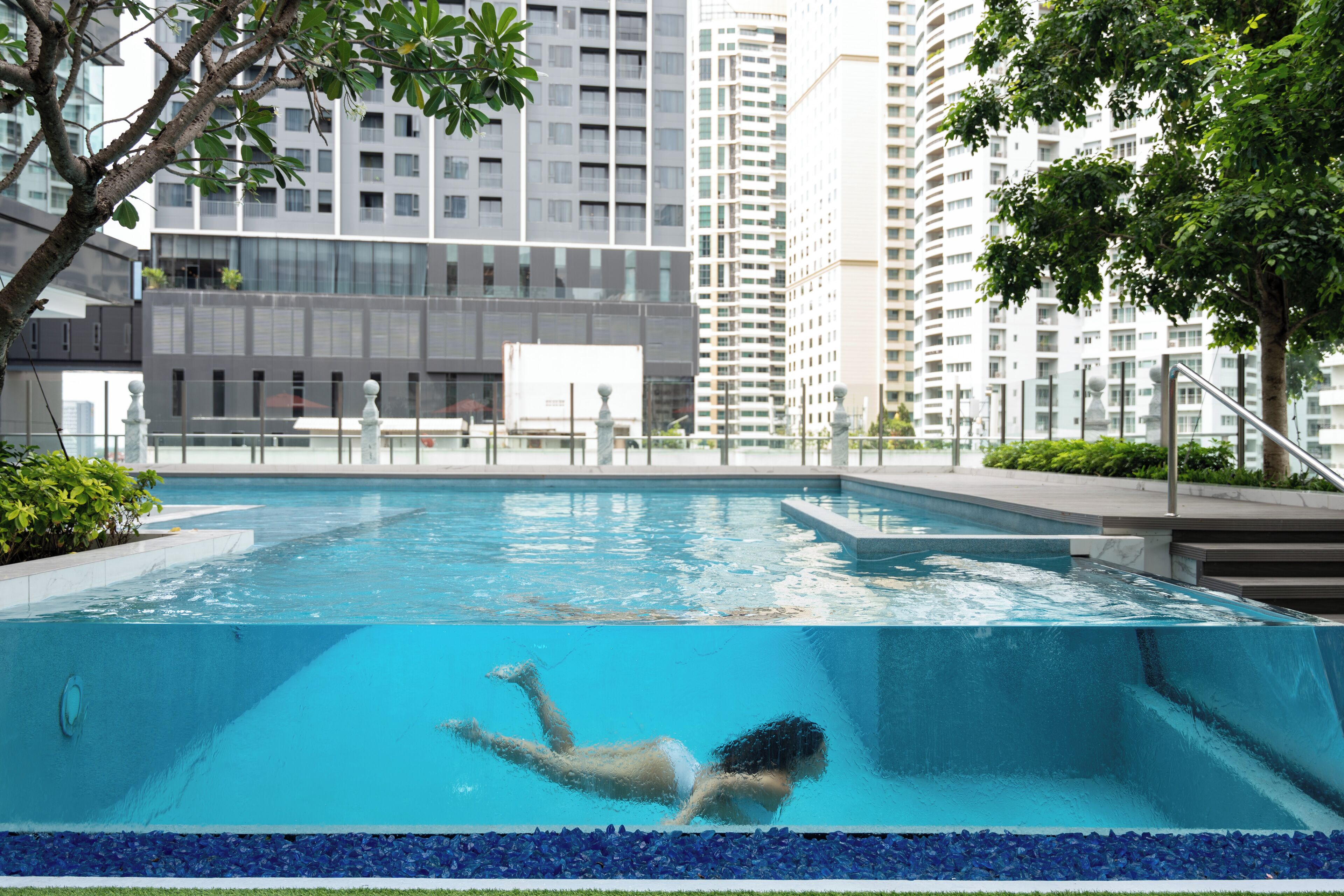 A swimming pool with a person swimming, surrounded by tall buildings and greenery.