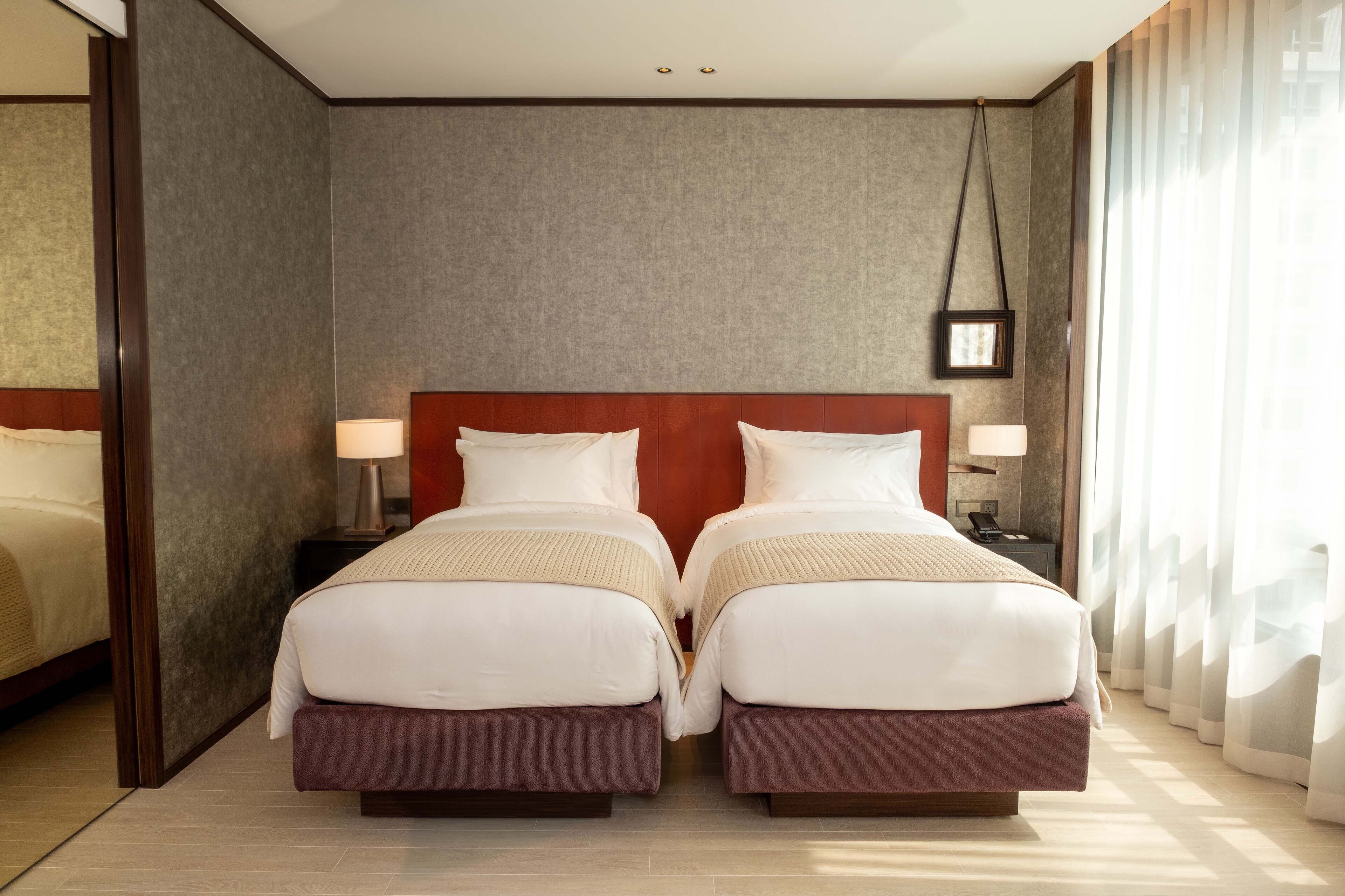 Een hotelkamer met twee bedden, een houten hoofdbord, nachtlampjes en een groot raam met dunne gordijnen.