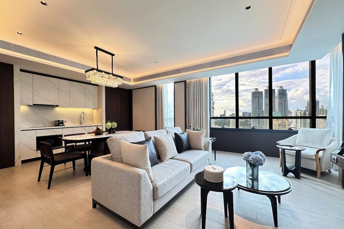 A modern living room with a sofa, dining table, and chairs, a kitchen area, and large windows offering a city view.