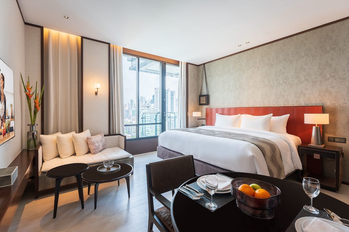 A hotel room with a large bed, a sofa, a dining table with a fruit basket and wine glasses, and a city view through large windows.