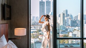 Habitación de hotel con un ventanal grande que ofrece vistas a la ciudad, una cama con almohadas y una lámpara de noche.