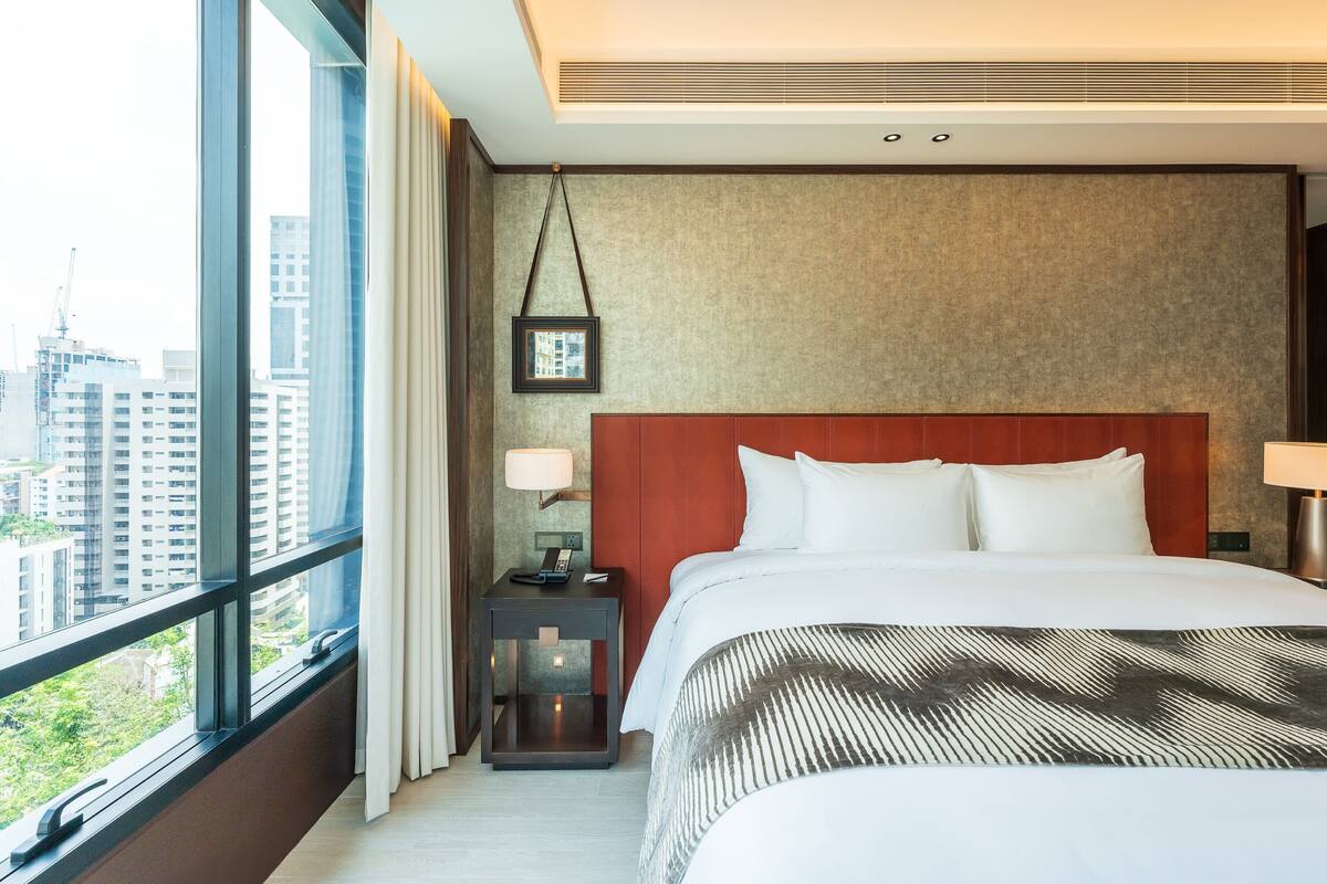A hotel room with a large window, a bed with white linens, a nightstand with a lamp, and a framed picture on the wall.