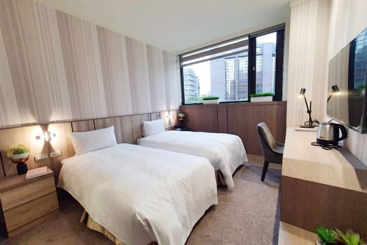 A hotel room with two beds, a desk, a chair, a TV, and a window with a city view.
