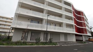 Un immeuble à plusieurs étages doté de balcons, d’un espace de stationnement et d’un élément vertical rouge distinctif sur le côté droit.