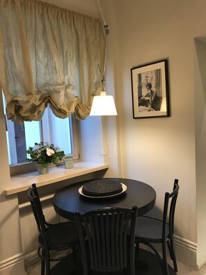 A dining area with a round table, black chairs, a window with curtains, a hanging lamp, and a framed picture on the wall.