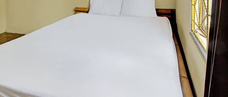 Una cama individual con ropa blanca en una habitación de paredes lisas y una ventana con marco decorativo.