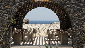 An outdoor dining area with stone walls and a view of the beach and ocean. - Elysian Retreat - Adults Only (Santorini)