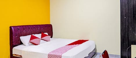 Un llit individual amb capçalera vermella i roba de llit blanca, una cadira i una taula petita en una habitació amb parets grogues i blanques.