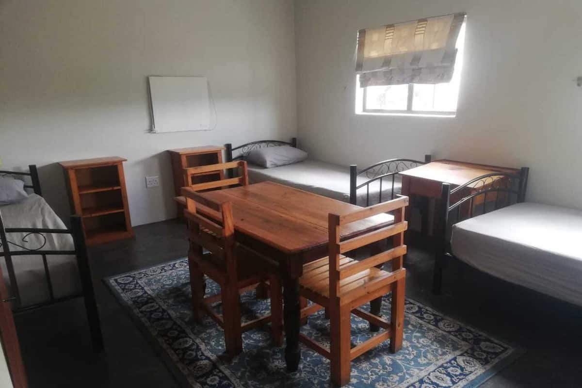 A small, compact room with a wooden table and chairs, a single bed, a small desk, and a window with curtains.