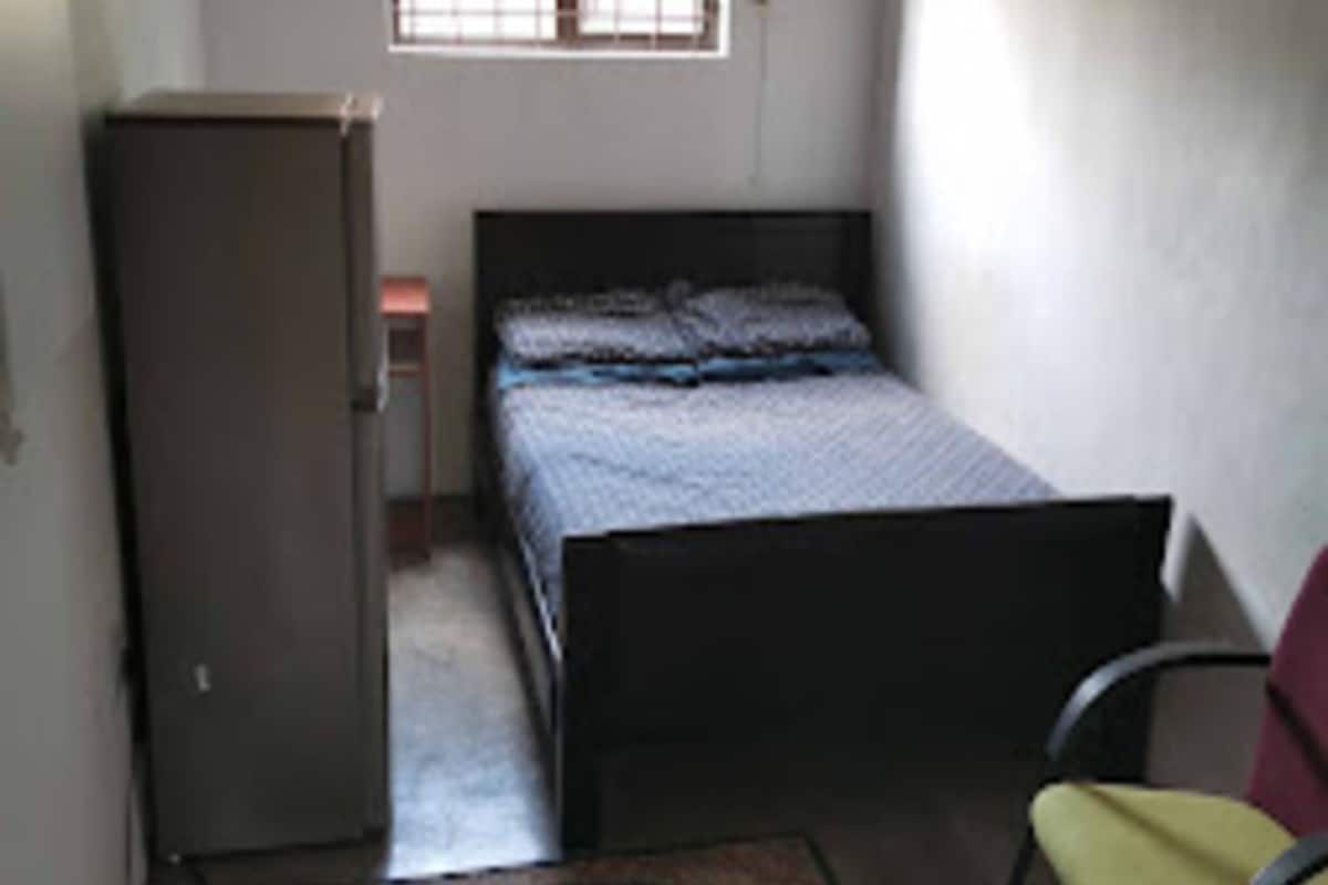 A small, dimly lit room with a single bed, a chair, a desk, and a window with a curtain.
