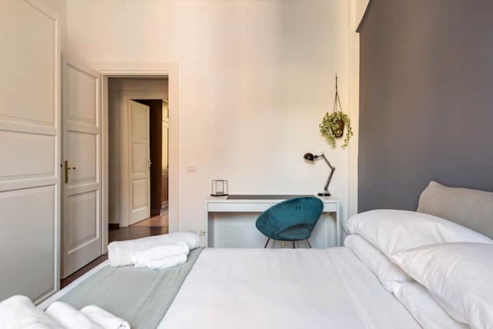 A hotel room with a bed, a desk with a chair, a hanging plant, and a door leading to another room.