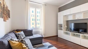 Un saló modern amb un sofà seccional gris, una televisió de pantalla plana sobre un moble blanc i una finestra gran amb vista a edificis.