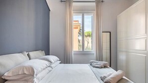 Un dormitori amb un llit, cortines blanques, una finestra i un armari.
