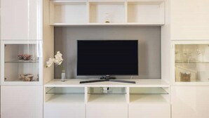 Un saló modern amb televisió de pantalla plana penjada en un moble blanc. Hi ha prestatges de vidre a ambdós costats de la televisió, i un ram de flors blanques a l'esquerra.