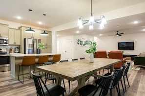 Una cocina moderna con electrodomésticos integrados, un comedor con mesa y sillas de madera, y una sala de estar con televisor y asientos.