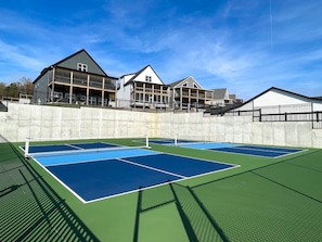 Una cancha de tenis al aire libre con una red, rodeada por un muro de concreto y un edificio con varios niveles y balcones.