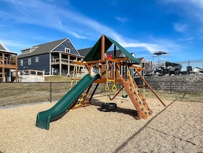 Un parque infantil con tobogán, red de escalada y columpios, rodeado por una valla y casas al fondo.