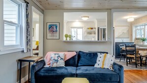 Ein Wohnzimmer mit einem dunkelblauen Sofa, einem gelben Hocker und einem Holztischchen. Im Hintergrund ist eine Küche mit weißen Schränken und einem Essbereich zu sehen.