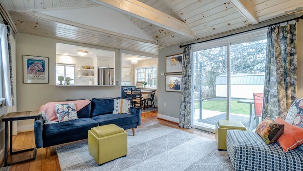 Ein großzügiges Wohnzimmer mit einem dunkelblauen Sofa, einem gelben Hocker und einem gemusterten Sessel. Im Hintergrund ist eine Küchenzeile erkennbar, und ein großes Schiebeglastür führt in einen Außenbereich.