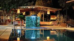 A nighttime pool area with a clear water surface reflecting lights, a stone structure, and a covered seating area. - Dar l'eau vive (Bine el Ouidane)