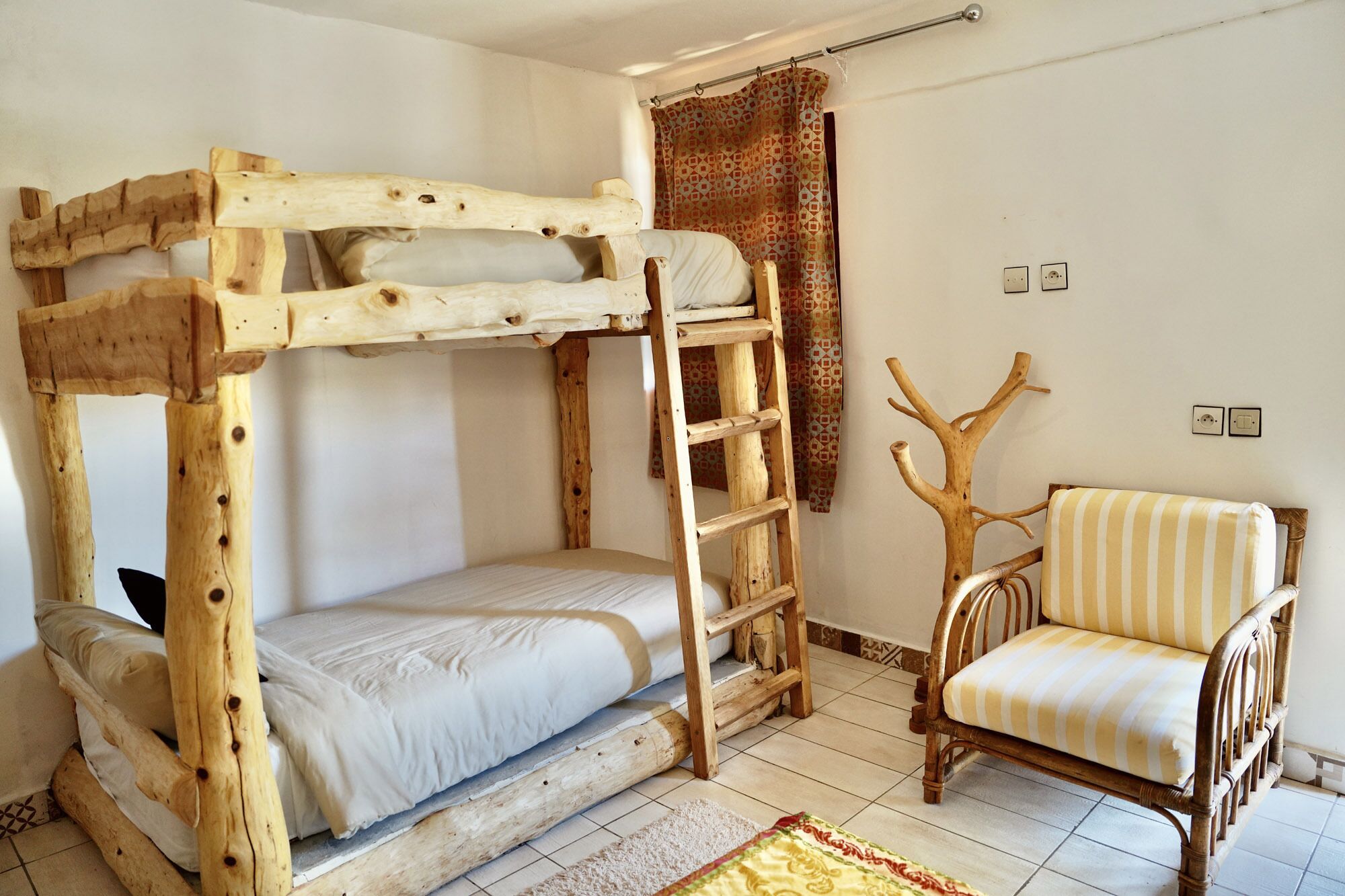 A bunk bed with a ladder, a wooden chair, and a patterned rug in a room with white walls and tiled flooring.