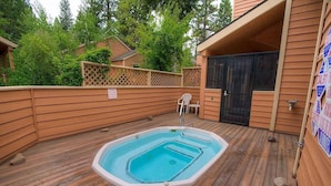 Une terrasse en bois avec un jacuzzi, un espace salon couvert et un bâtiment doté d’une porte noire.