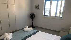 A bedroom with a bed, a standing fan, a window with blinds, and a small bedside table. - Freshly Refurbished Apartment in Paralimni, Cyprus (Paralimni)