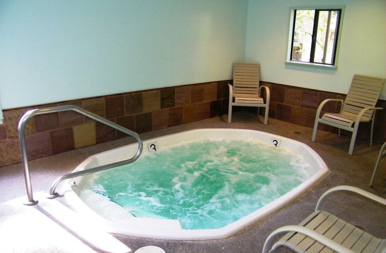 A hot tub with a metal ladder, surrounded by chairs, in a room with tiled walls and a window.