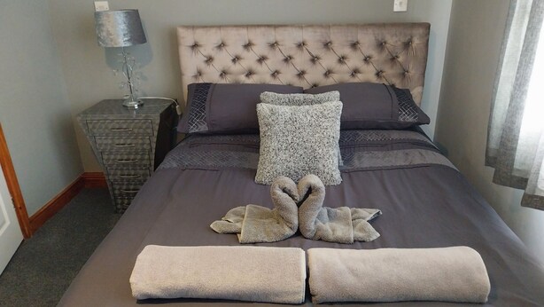 A neatly made bed with a tufted headboard, a bedside table with a lamp, and a dresser. - 3-bed Apartment in Lowestoft With sea Views (Lowestoft)